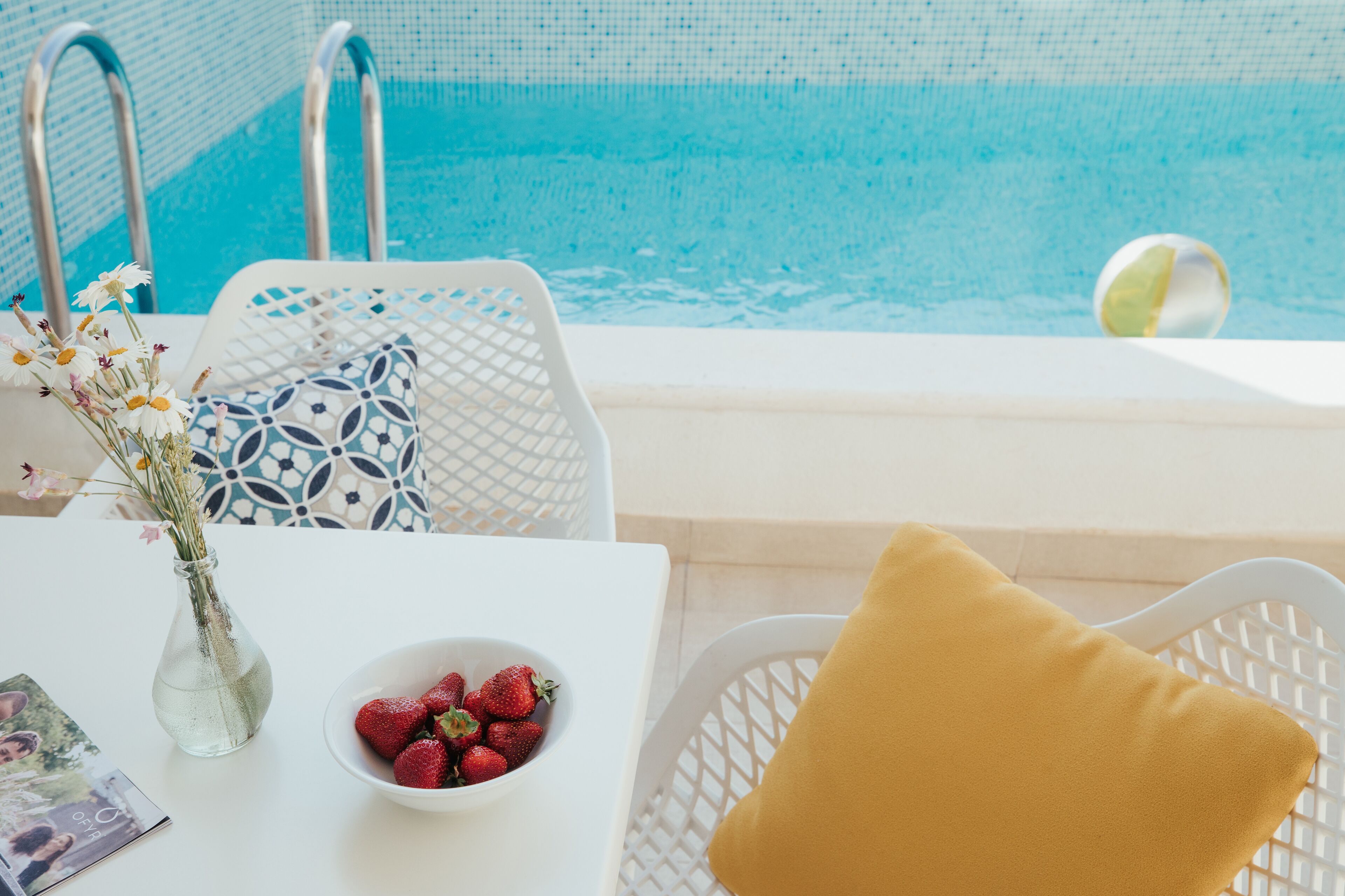 Zona de piscina con una mesa blanca, un cuenco de fresas, un jarrón con flores y una almohada amarilla.