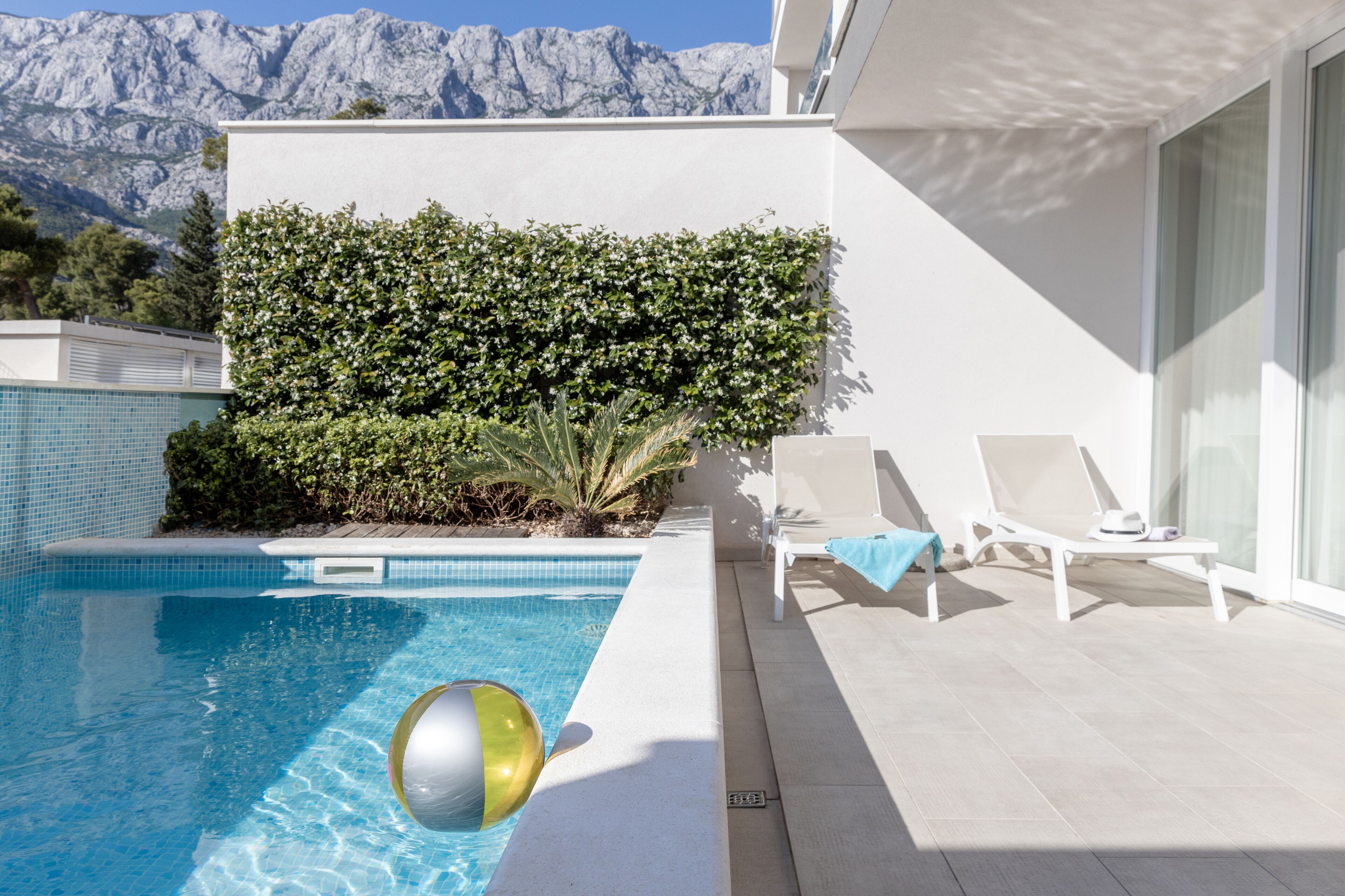 Une piscine avec une vue sur les montagnes, des chaises longues blanches et une haie.