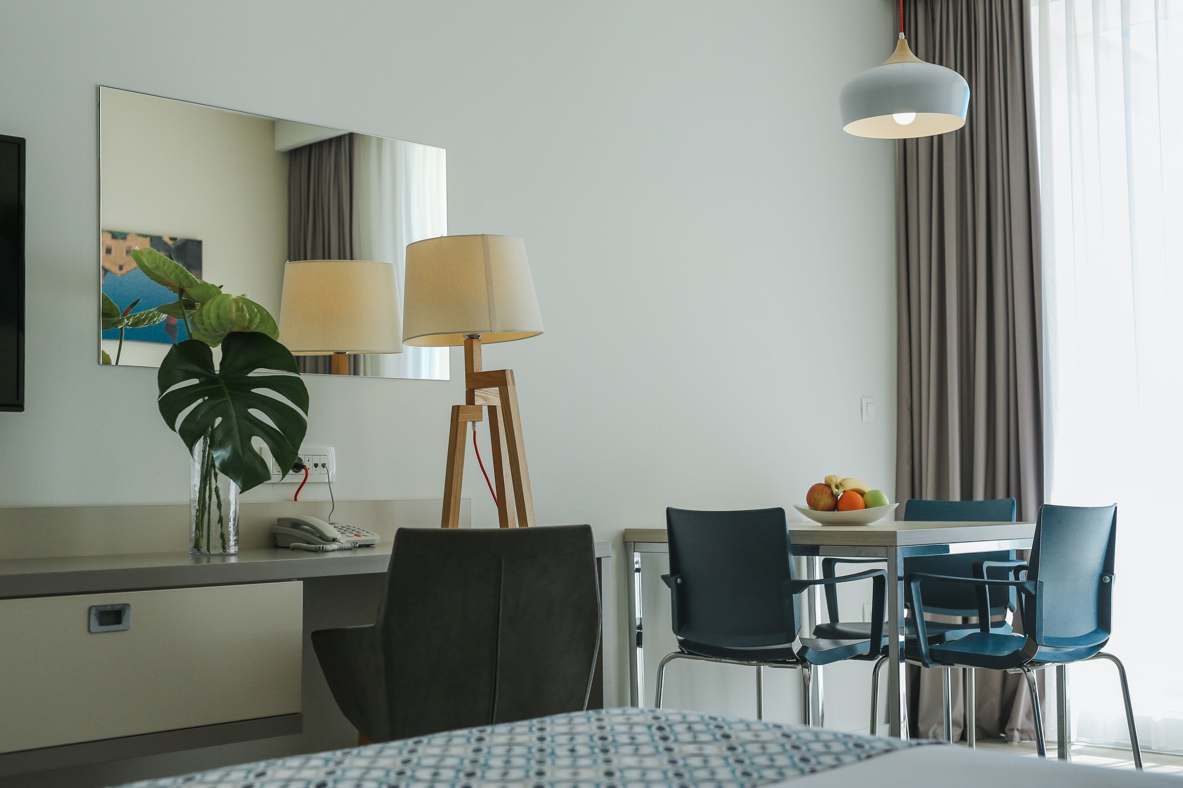 A hotel room with a bed, a desk with a lamp, a chair, a table with fruit, and a large window with curtains.