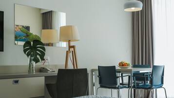 A hotel room with a bed, a desk with a lamp, a chair, a table with fruit, and a large window with curtains.