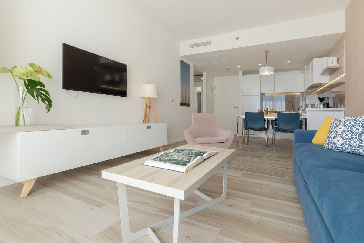 Een moderne woonkamer met een flatscreen televisie, een witte bank, een blauwe zithoek en een eethoek met stoelen en een tafel.