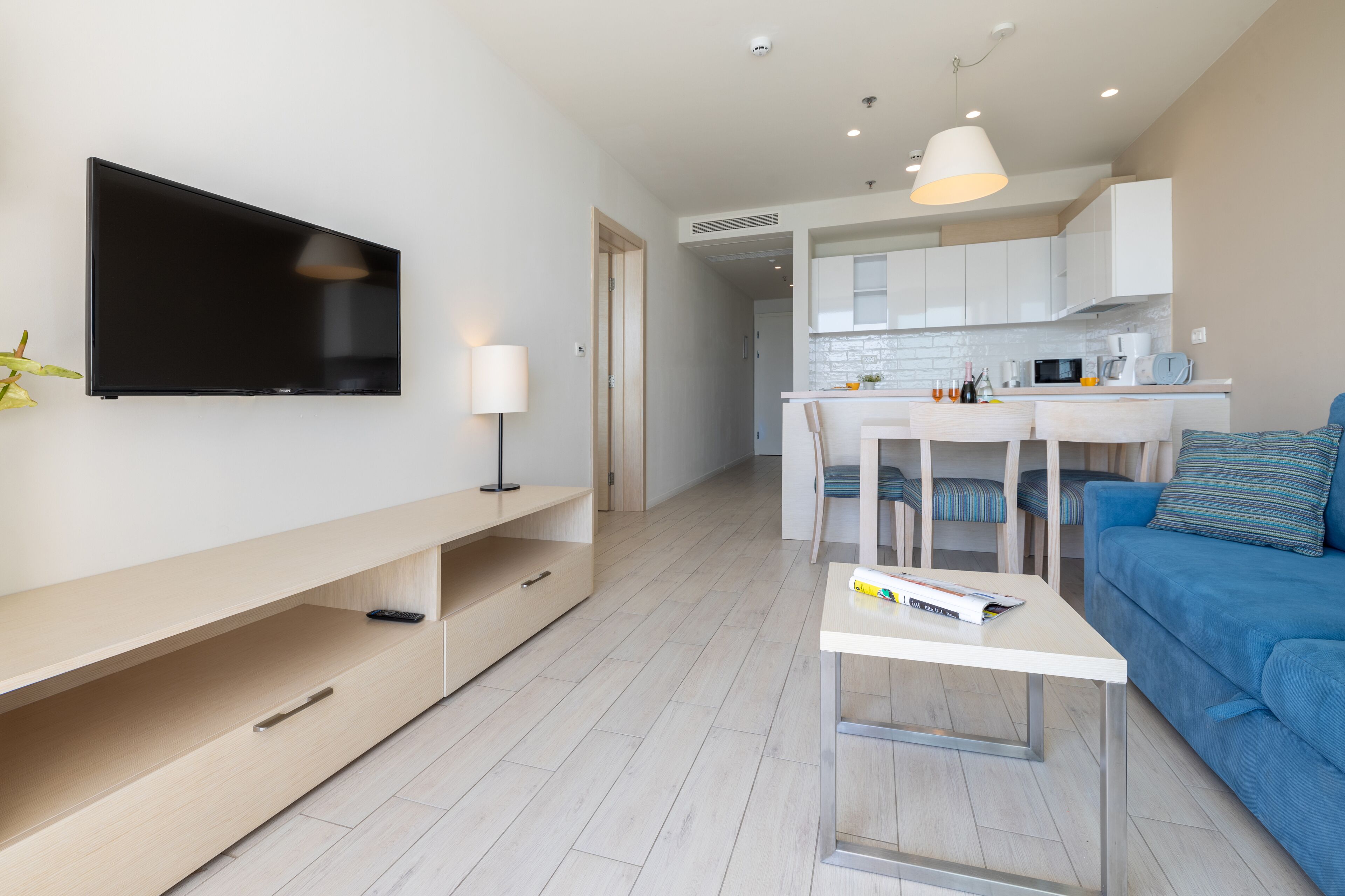 A modern living room with a flat-screen TV, a coffee table, a sofa, and a dining area with a kitchen in the background.