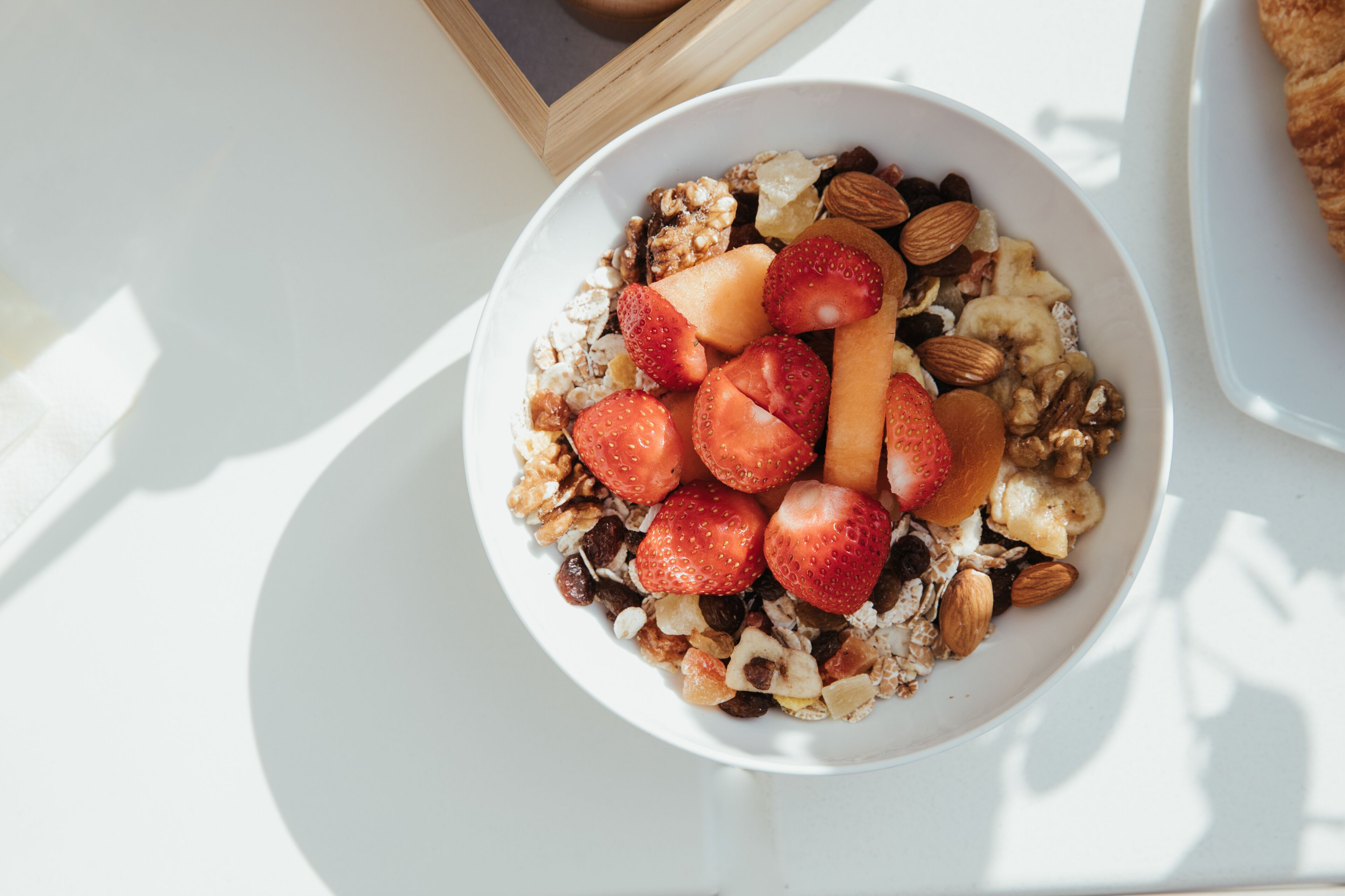 Un tazón de cereal con fresas, almendras y otras frutas sobre una mesa blanca.