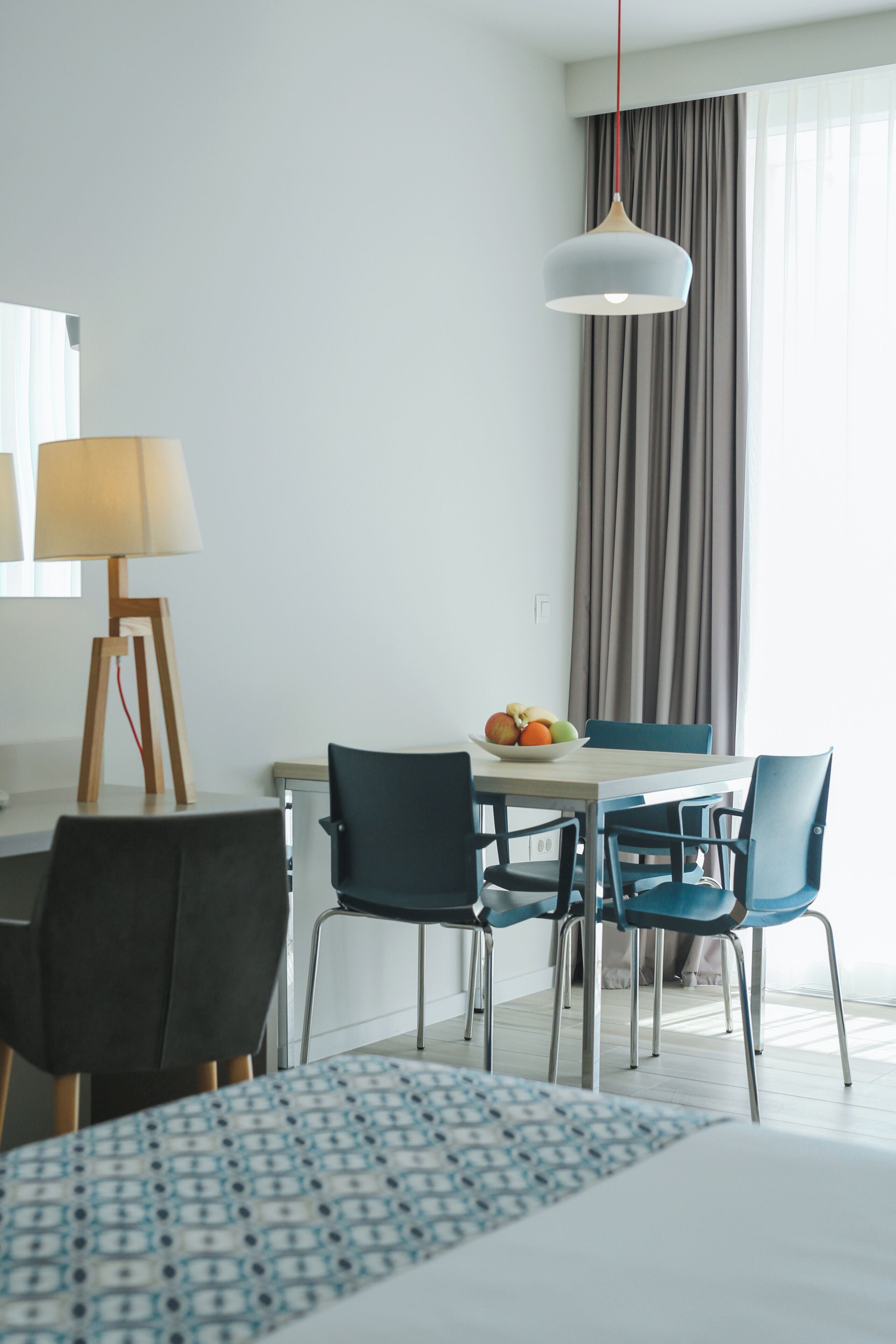 A hotel room with a bed, a desk with a lamp, a dining table with chairs, and a fruit bowl.