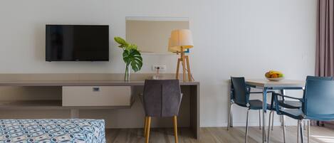 Quarto de hotel com cama, secretária com computador, cadeira, mesa de jantar com cadeiras, luminária e televisão na parede.
