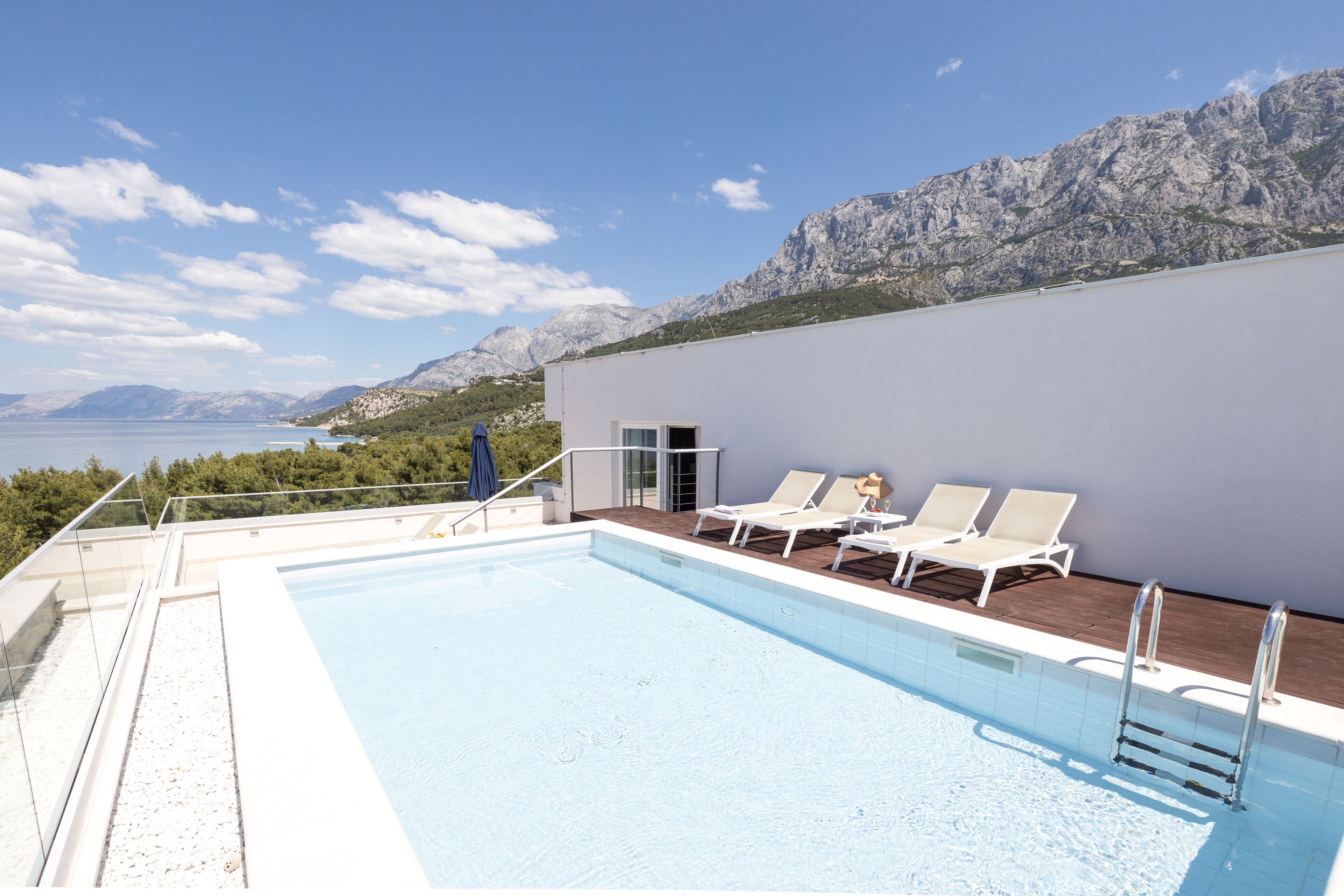 A rooftop pool with white lounge chairs and a glass balustrade, overlooking a mountainous landscape and a body of water.