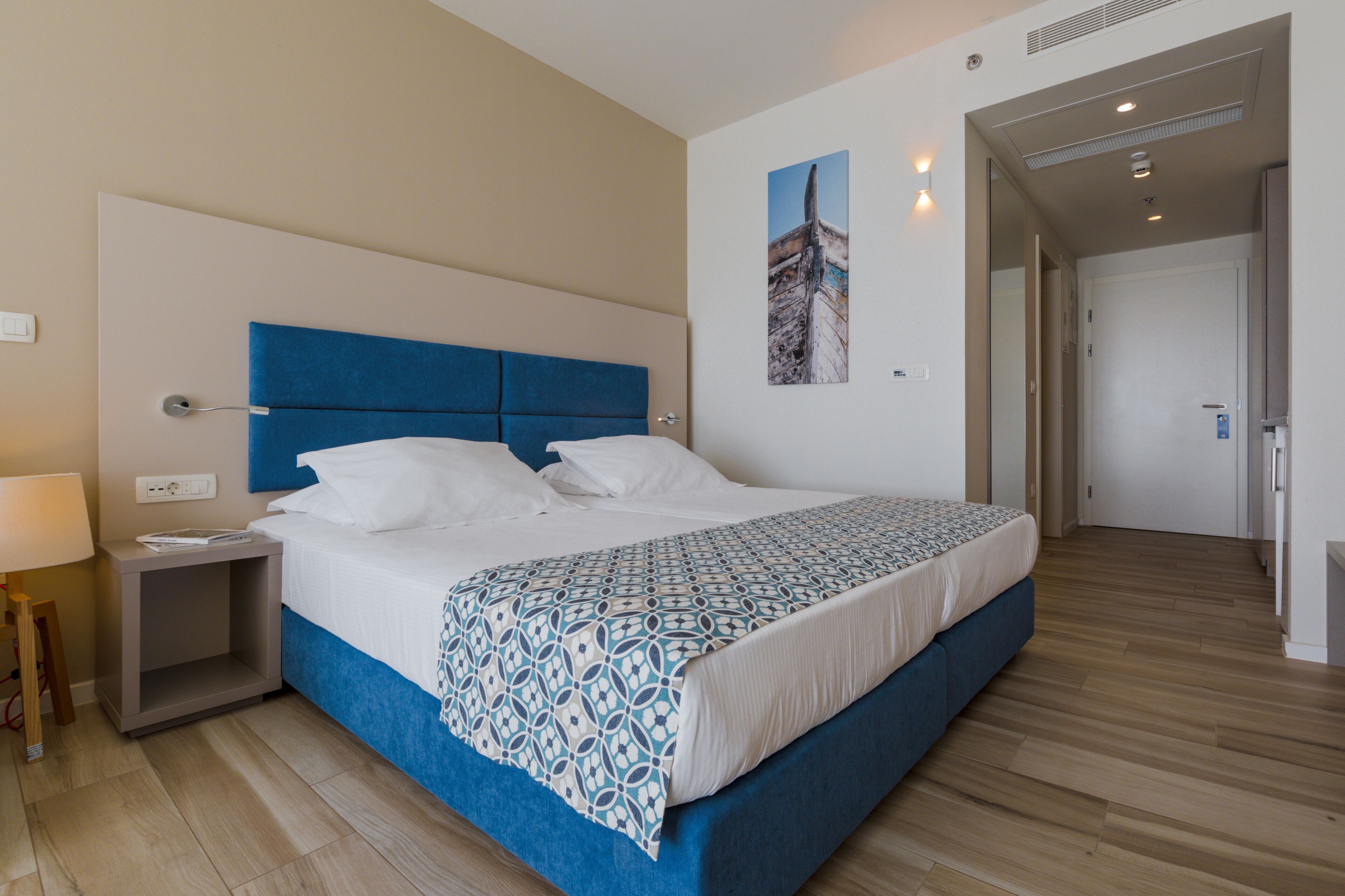 A hotel room with a large bed, a bedside table, a lamp, a wall-mounted power outlet, and a door leading to another room.