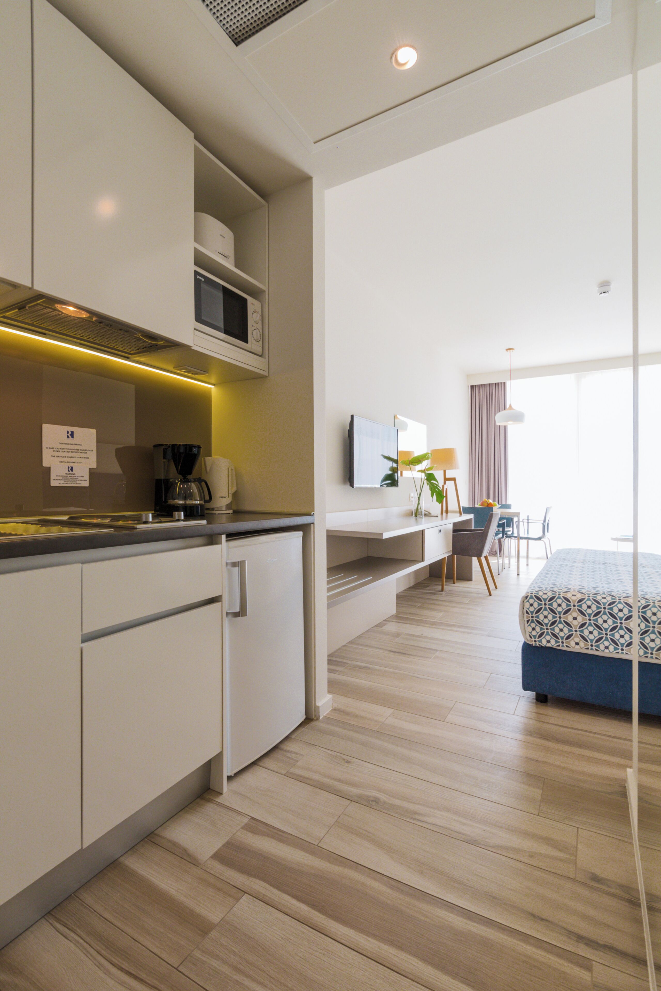 Una habitación de hotel moderna con kitchenette, una cama con ropa de cama estampada, un escritorio con silla y un gran ventanal que proporciona luz natural.