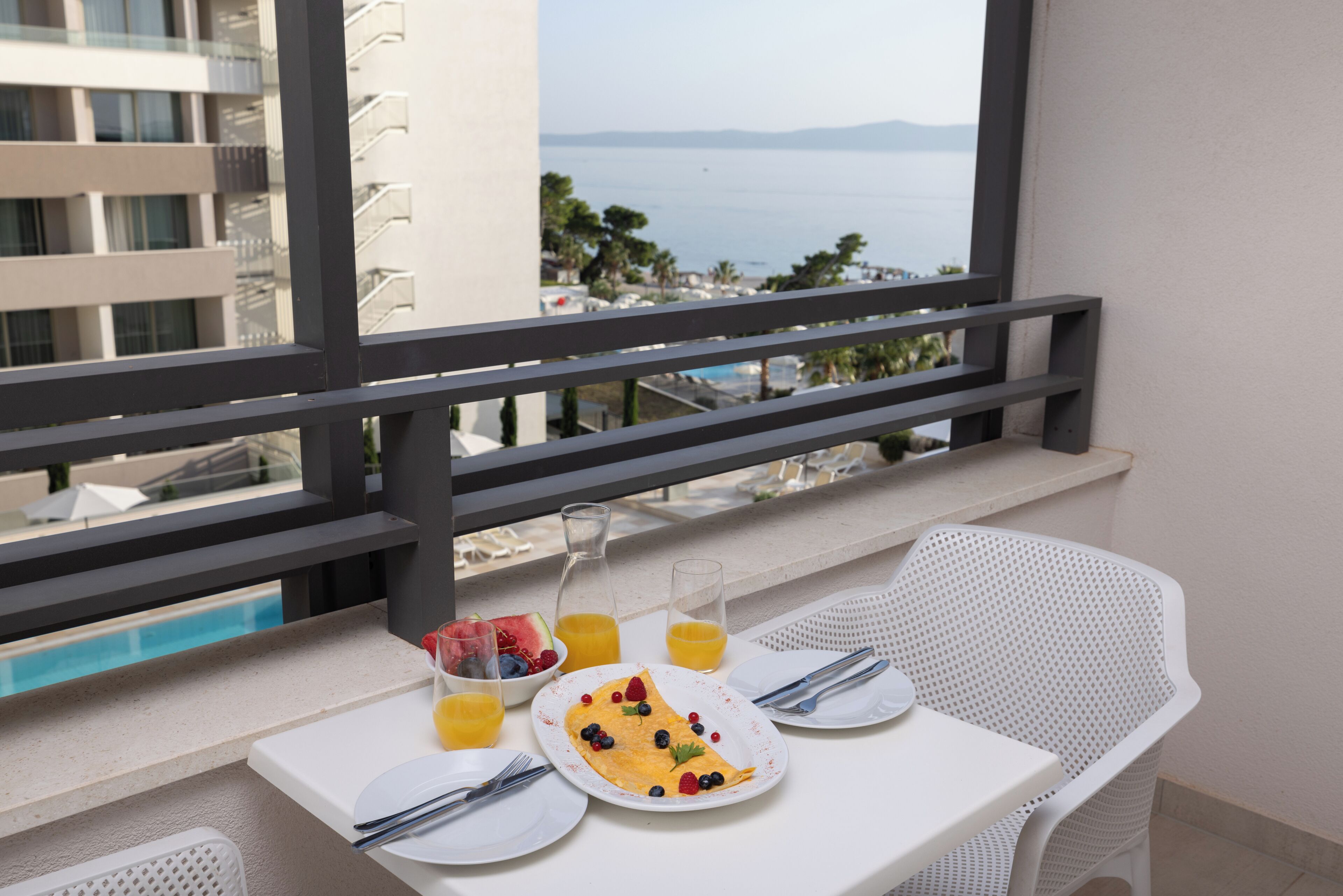 A balcony with a table set for breakfast, a view of the sea, and a pool in the background.