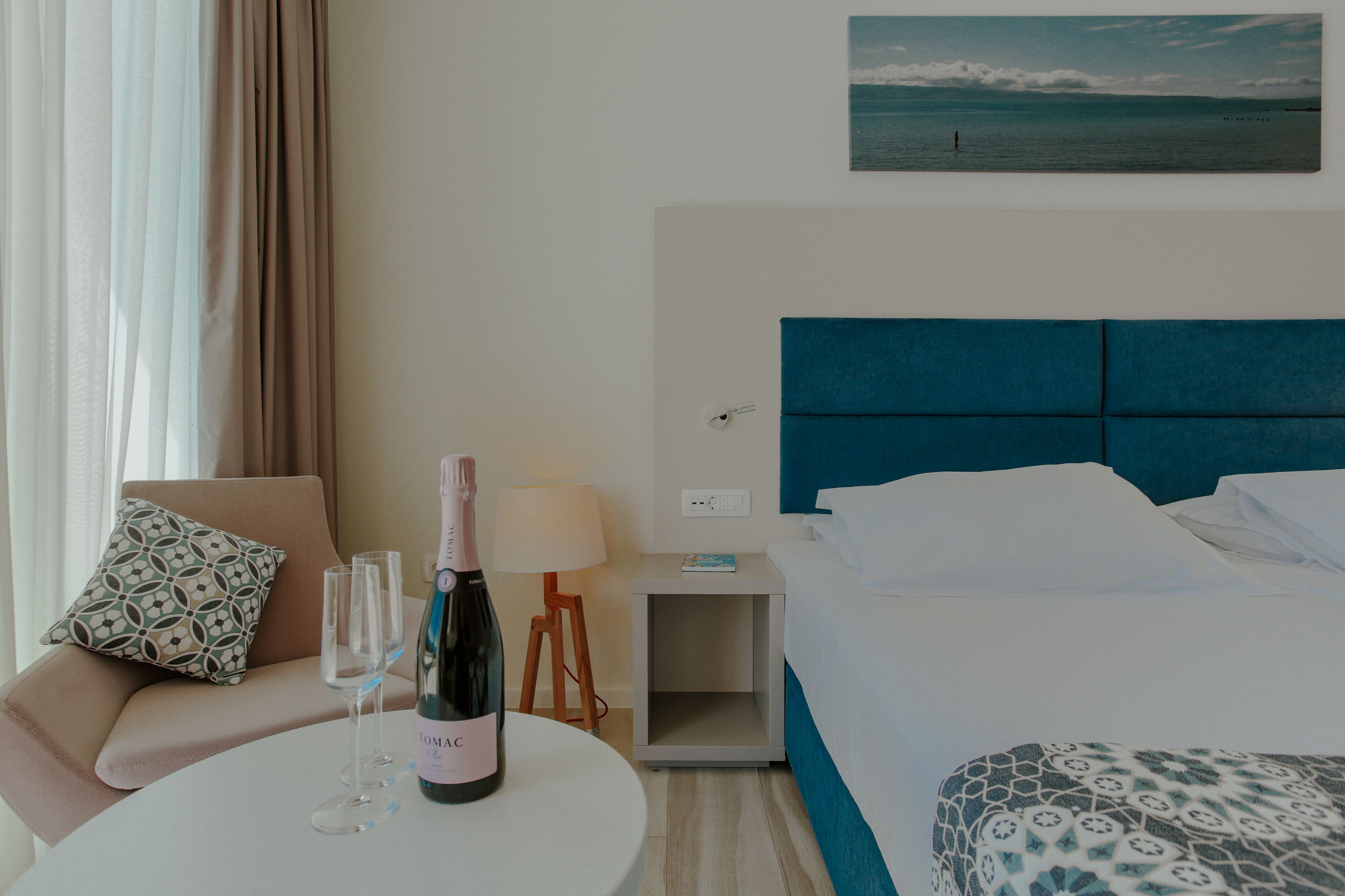 A hotel room with a bed, a bedside table with a lamp, a chair with a patterned cushion, a small table with a bottle of champagne and glasses, and a painting of the sea on the wall.