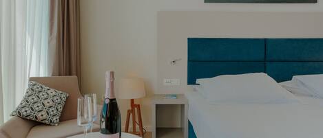 A hotel room with a bed, a bedside table with a lamp, a chair with a patterned cushion, a small table with a bottle of champagne and glasses, and a painting of the sea on the wall.