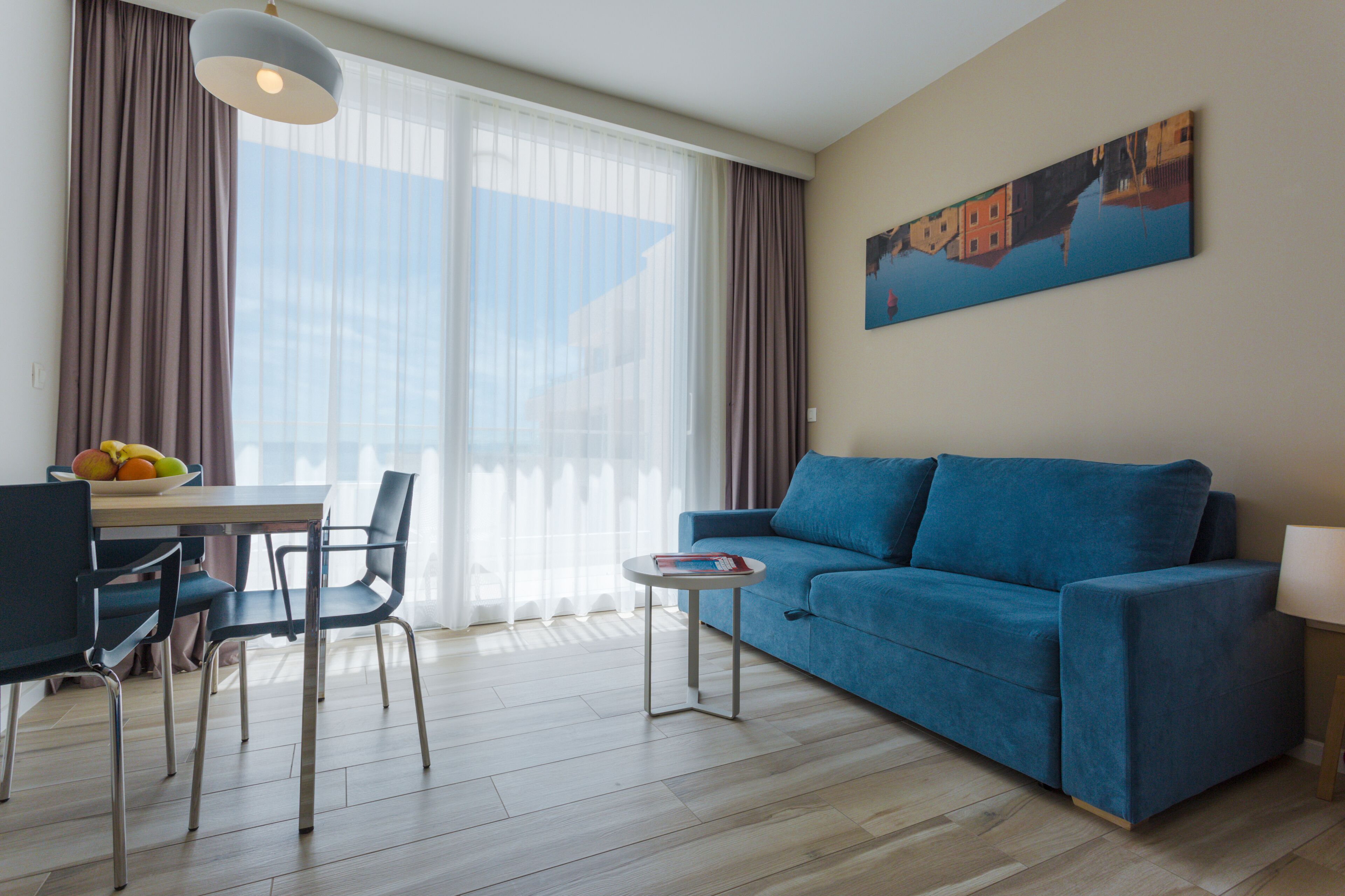 Een moderne woonkamer met een blauwe bank, een eettafel met stoelen, een klein rond tafeltje met een boek, en een groot raam met gordijnen.