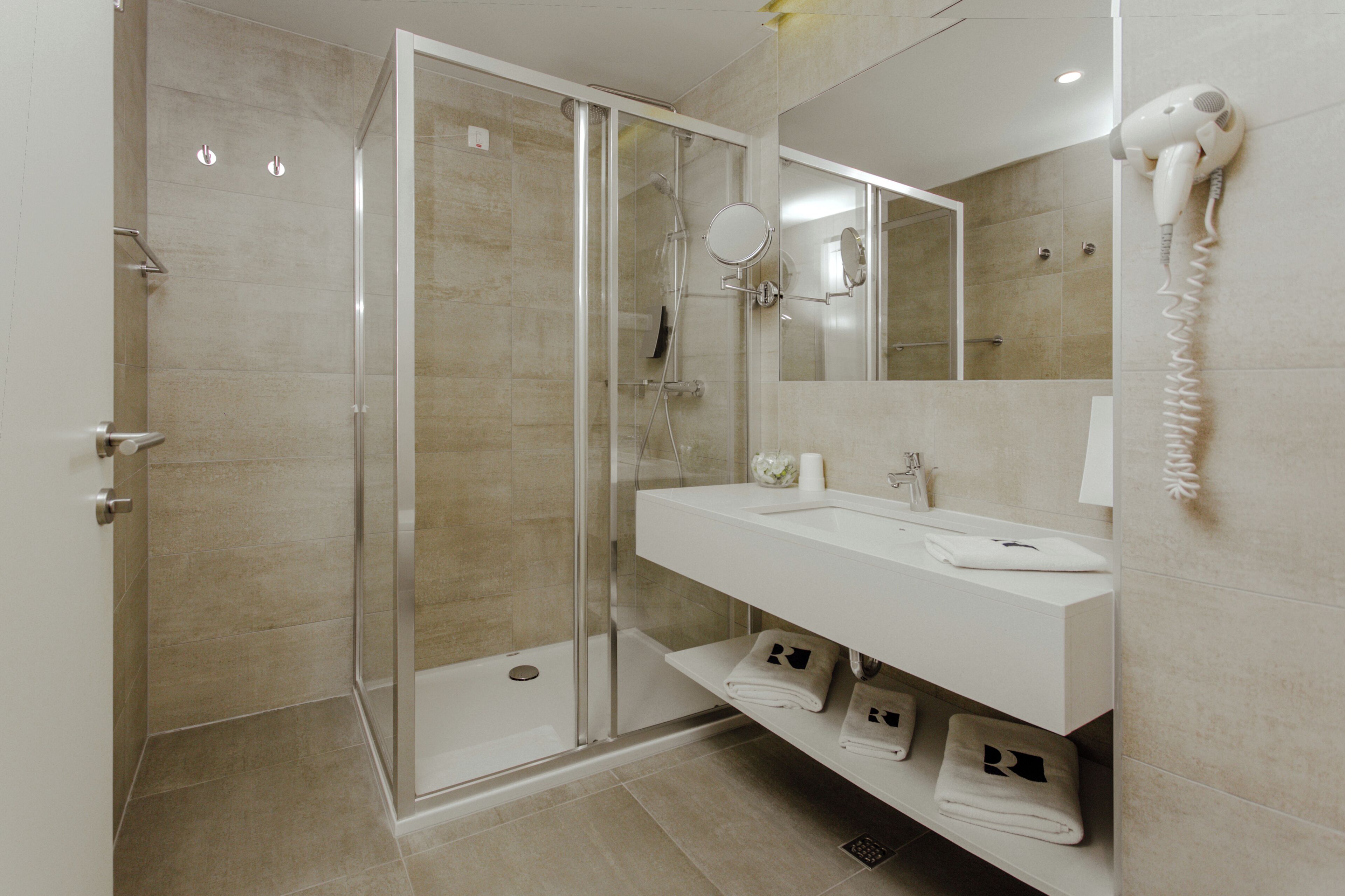 A modern bathroom with a glass-enclosed shower, a white sink, and a hairdryer mounted on the wall.