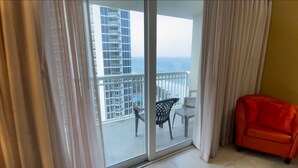 Een kamer met een schuifdeur naar een balkon met uitzicht op een hoog gebouw en de oceaan. Er staat een klein tafeltje en twee stoelen op het balkon.