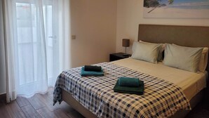 Un dormitorio con colcha a cuadros, dos almohadas, una mesita de noche con lámpara, un cuadro con una playa y una ventana con cortinas traslúcidas.