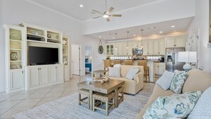 Ein großzügiges Wohnzimmer mit Fernseher, Sofa und Couchtisch, eine Küche mit modernen Geräten und ein Deckenventilator.