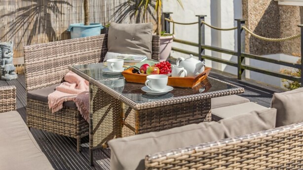 Un coin salon extérieur aménagé avec des meubles en osier, une table en verre, des théières blanches, des tasses et un saladier rempli de fruits.