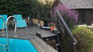 Una terraza de madera con piscina, sillas de descanso y un cobertizo pequeño con flores moradas.