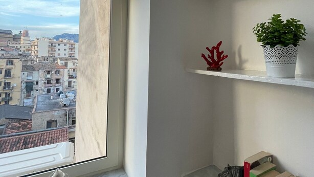 A room with a large window overlooking a cityscape, a white radiator, a shelf with decorative items, and a small plant. - Luxury Apartament's Celso (Palermo)