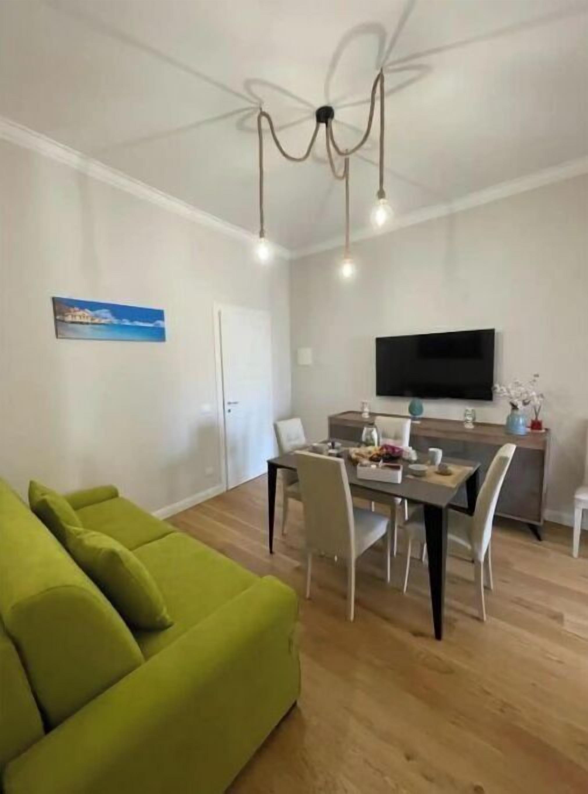 Un salon moderne avec un canapé vert, un coin repas comprenant une table et des chaises, un téléviseur à écran plat fixé au mur et un lustre à trois lampes.
