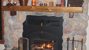 Une cheminée en pierre avec un feu, une étagère en bois au-dessus et un pot en cuivre posé sur le sol.