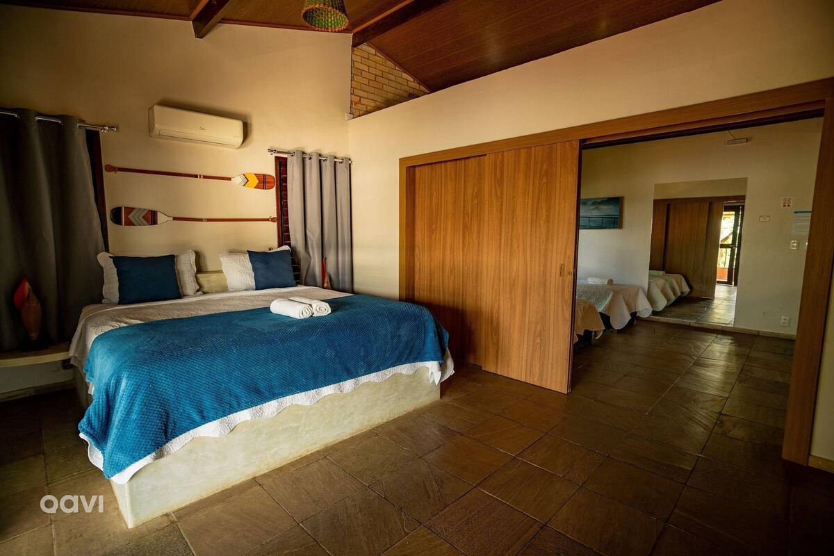 A bedroom with a bed, an air conditioner, a wooden door, and a view into another room.