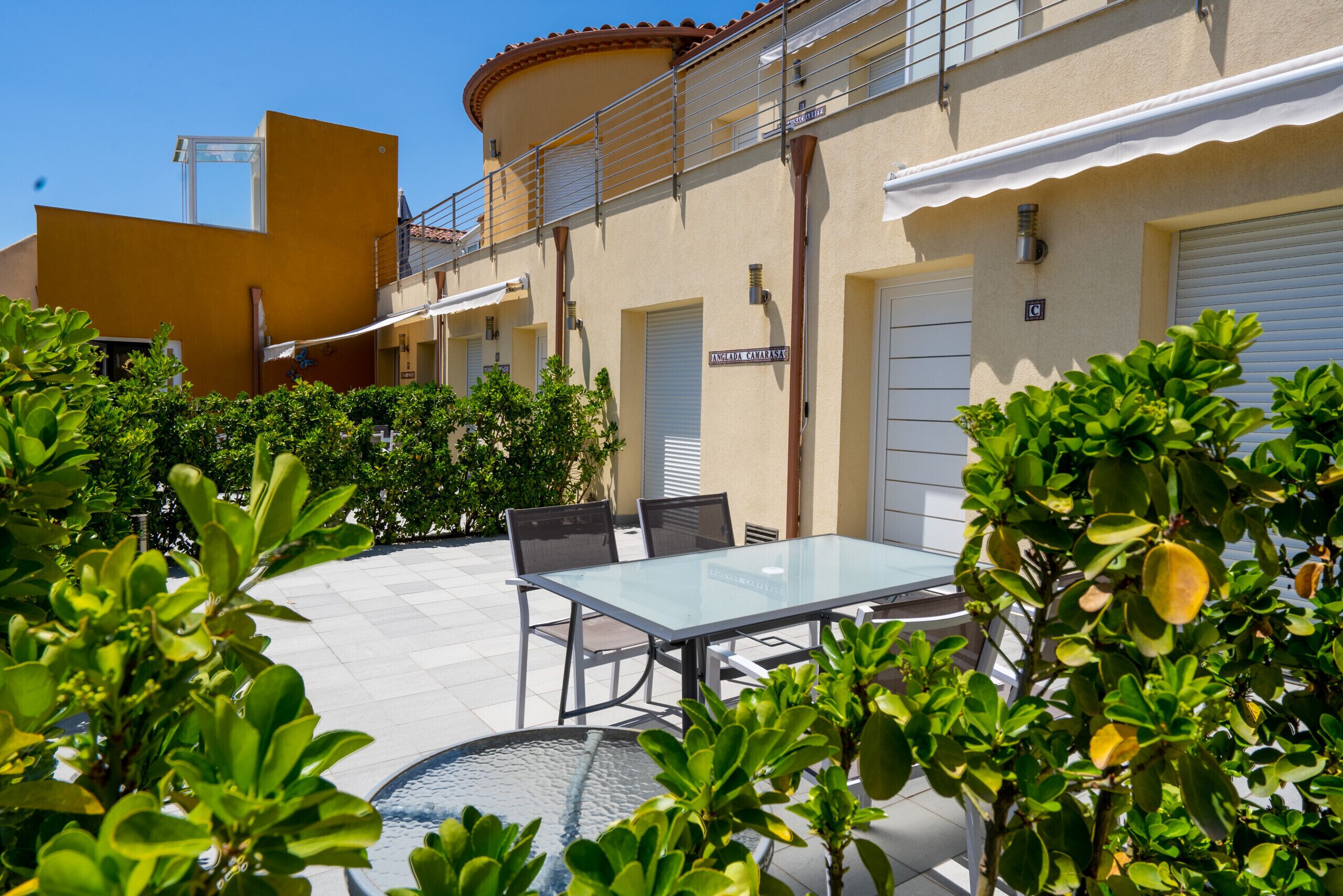 Un patio con una mesa y sillas de vidrio, rodeado de follaje y un edificio de fachada color beige y tejado de tejas rojas.