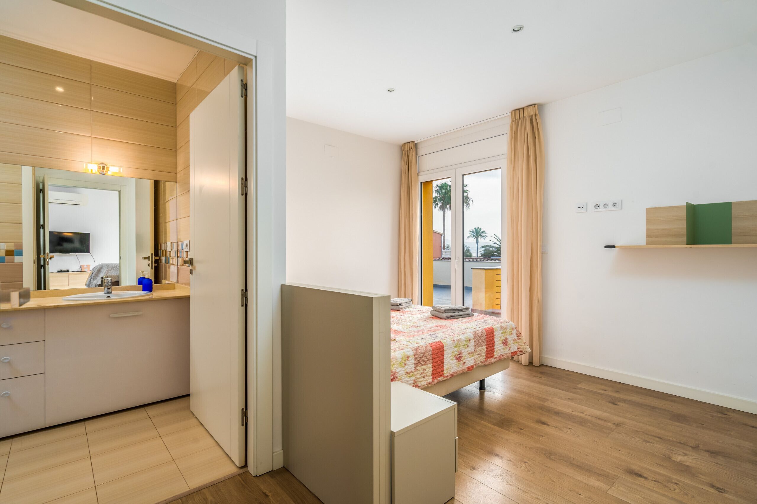 A modern bedroom with a wooden floor, a bed with a floral bedspread, a bedside table, a nightstand, a mirror, a wardrobe, and a view of the outside through a window.