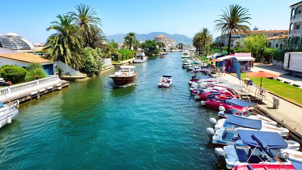 Un port de plaisance où des bateaux sont amarrés le long de la rive, un pont et des bâtiments dotés de balcons.