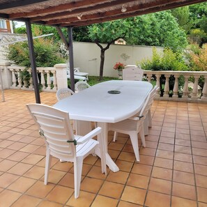 Un patio couvert avec une table et des chaises blanches, entouré d’un sol carrelé et d’une rambarde décorative.