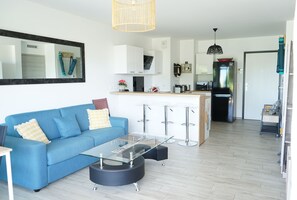 A modern living room with a blue sofa, a glass coffee table, and a kitchen area with white cabinets and a black refrigerator.