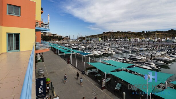 Un puerto deportivo con numerosas embarcaciones amarradas, un paseo peatonal y un edificio colorido con balcones.