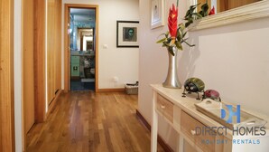 Un pasillo con piso de madera, una pared blanca con un cuadro enmarcado, un florero con flores y un armario de madera con espejo.