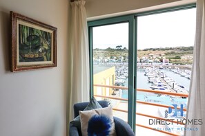 Ein Zimmer mit großem Fenster und Blick auf eine Marina mit Booten, einem gerahmten Gemälde an der Wand und einem Sessel mit Kissen.