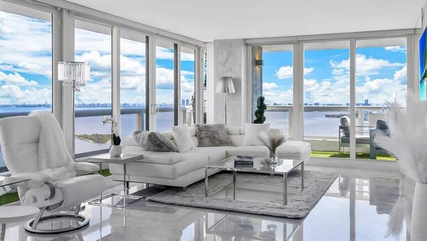 Uma sala de estar moderna com um sofá branco, uma mesa de centro de vidro e vista para a água através de grandes janelas.