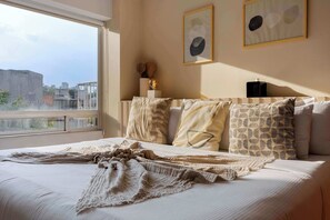 A bedroom with a large window, a bed with patterned pillows, and two framed artworks on the wall.