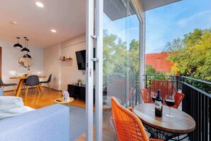 A modern living room with a dining area, a balcony with outdoor seating, and a large window offering a view of trees and buildings.