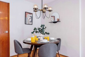 A dining area with a round table, grey chairs, a chandelier, a potted plant, and a wall-mounted shelf with a mirror.