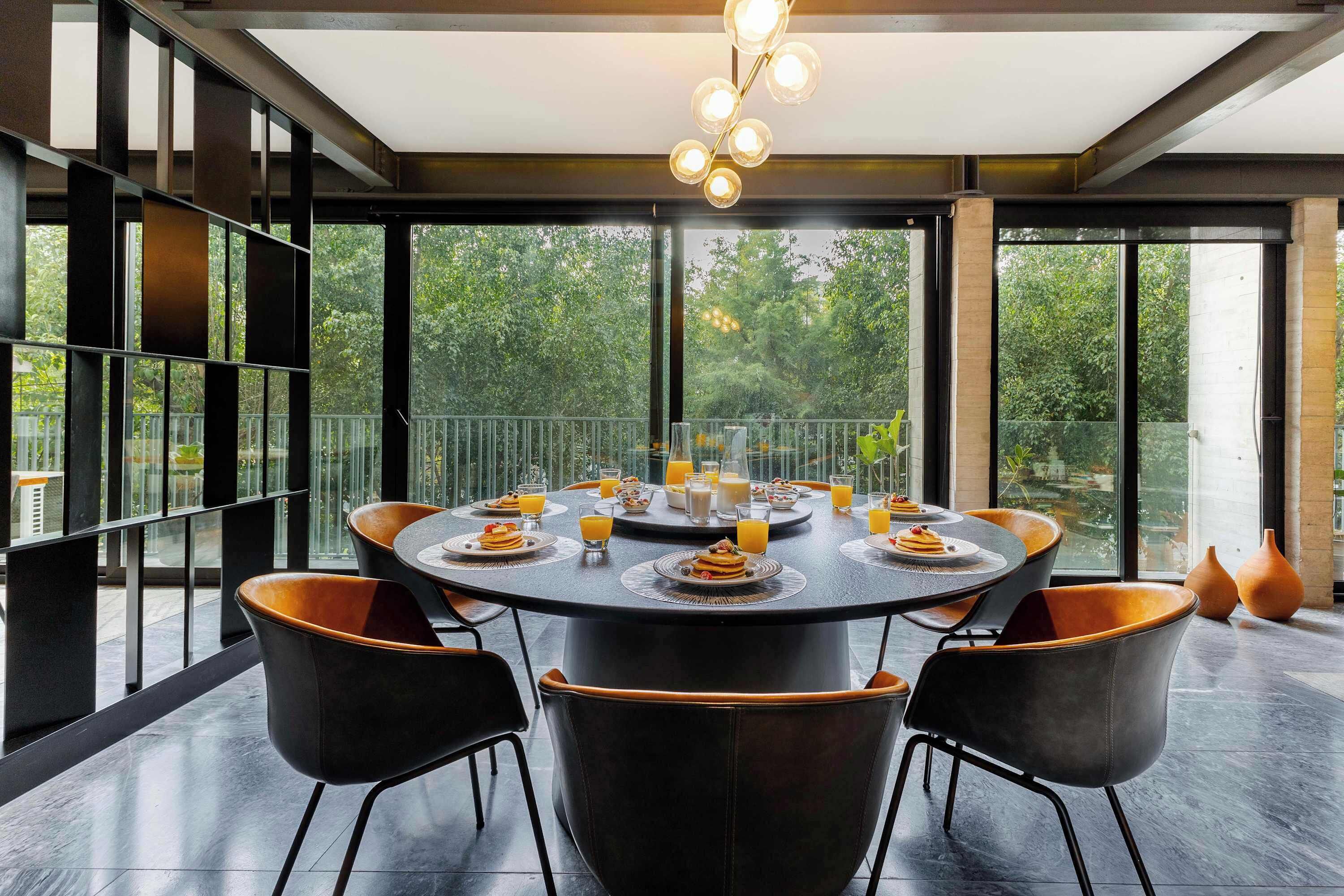 A modern dining area with a round table, black chairs, and a view of greenery through large windows.