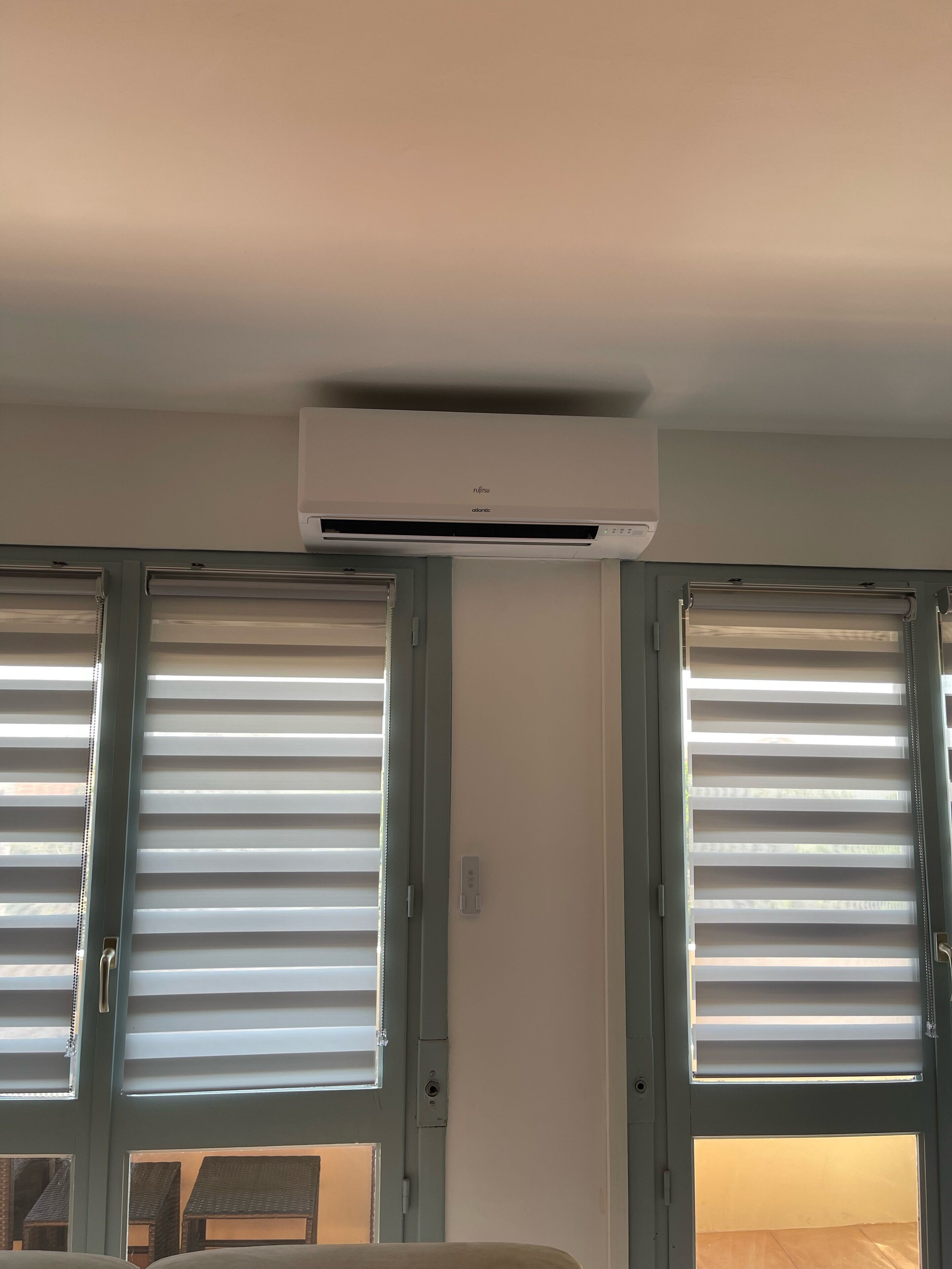 A room with an air conditioning unit mounted on the wall, a window with blinds, and a partial view of a bed.