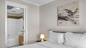 A bedroom with a bed, a nightstand with a lamp, a bathroom with a sink, and a painting on the wall.