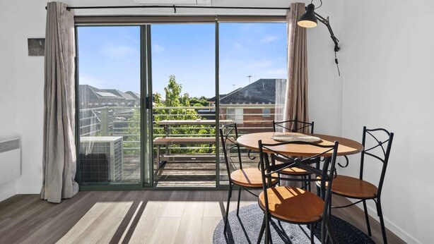 A room with a sliding glass door leading to a balcony, a round wooden table with four chairs, a black desk lamp, and a view of a cityscape and greenery.