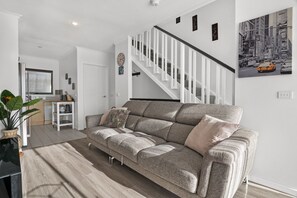 A modern living room with a grey sofa, a staircase, and a cityscape painting. - Snug 2-bed Townhouse Near Bustling Shops (Maribyrnong)