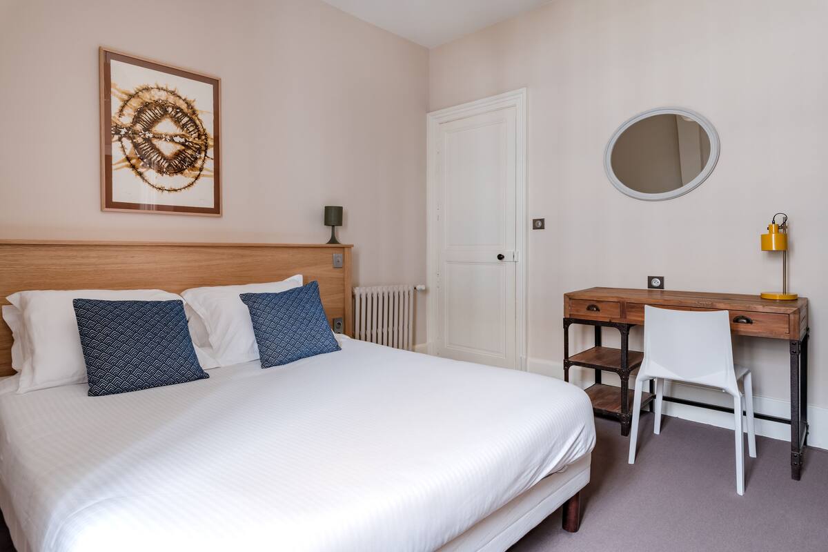 A neatly arranged bedroom with a bed, a desk, a chair, a mirror, and a framed artwork on the wall.