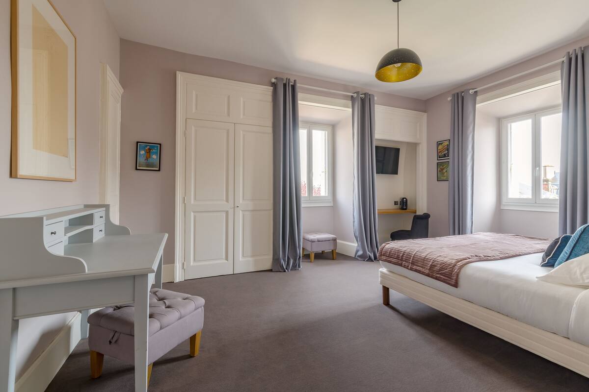 A bedroom with a bed, a desk, a chair, and a small stool. There are curtains on the windows and a picture on the wall.