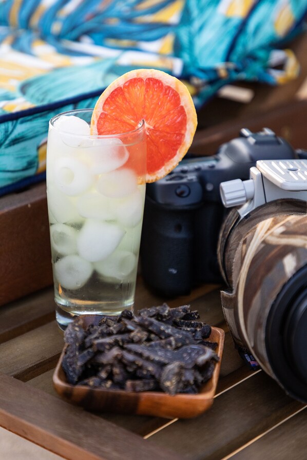Un vaso con cubitos de hielo y una rodaja de naranja, una cámara, y un cuenco de fruta seca sobre una superficie de madera.