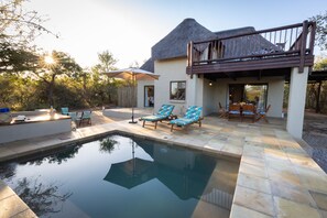 Un área de piscina con sillones de descanso y una mesa de comedor, rodeada por un deck de piedra y un edificio con techo de paja.