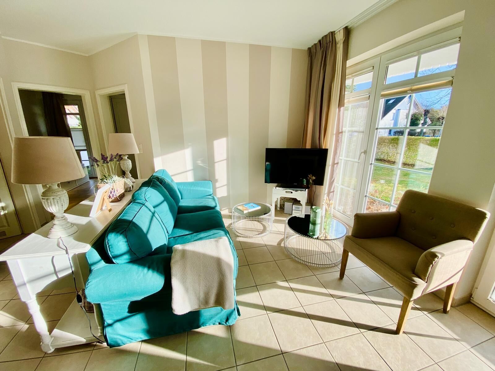 A living room with a teal sofa, a beige armchair, a TV, and a window with curtains.