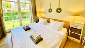 Ein Hotelzimmer mit einem großen Bett, zwei Kissen, einem Nachttisch mit Lampe und Blick auf einen Garten durch ein großes Fenster.