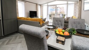 A modern living room with a sofa, dining table, and chairs. There is a television on a stand and a window with a view of mountains. - Red Kite Lodge (Crieff)